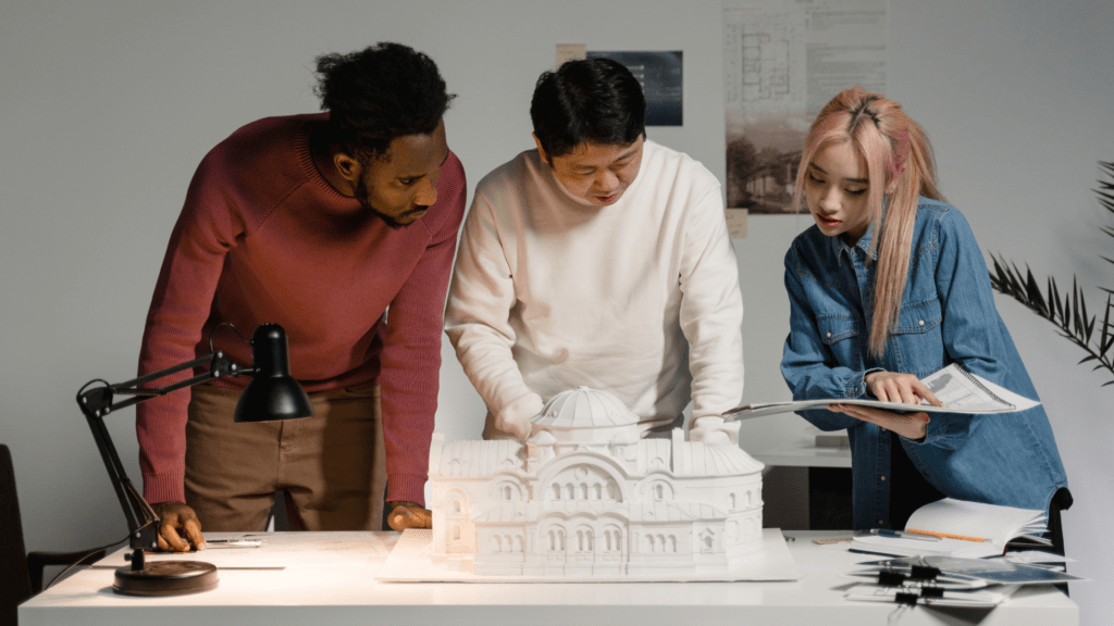 Three adult students are collaborating on a project in a modern workspace. They are gathered around a table with a detailed scale model of a building. One man in a red sweater leans in to examine the model, while another man in a white sweater points at it. A woman with pink hair, wearing a denim shirt, looks at design plans in a book. The table is well-lit with a desk lamp, and the atmosphere is focused and collaborative.