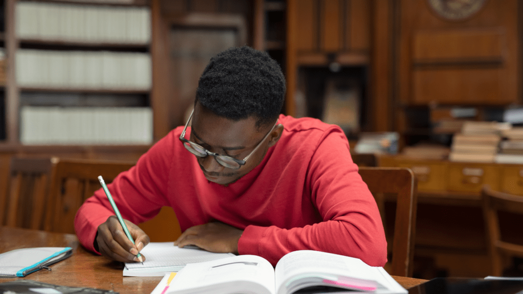 A student in a red shirt diligently writing in a book while studying alone in a library setting.
