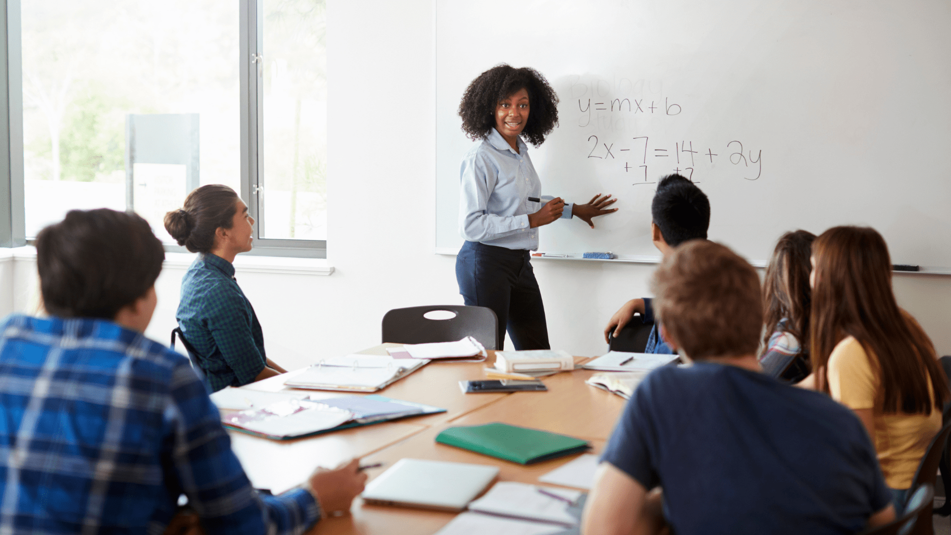 A teacher standing at a whiteboard, writing a math equation while a group of students seated at a table attentively watches and takes notes. The classroom setting indicates an interactive learning environment focused on solving problems together.