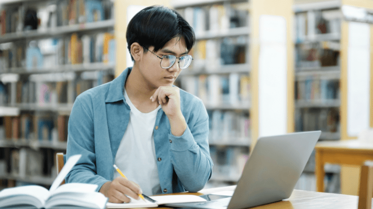 A student studying alone in a library, focused on a laptop screen while taking notes with a pencil. Open books are spread out on the table, indicating an intense study or research session. The background shows rows of bookshelves, emphasizing the academic setting.