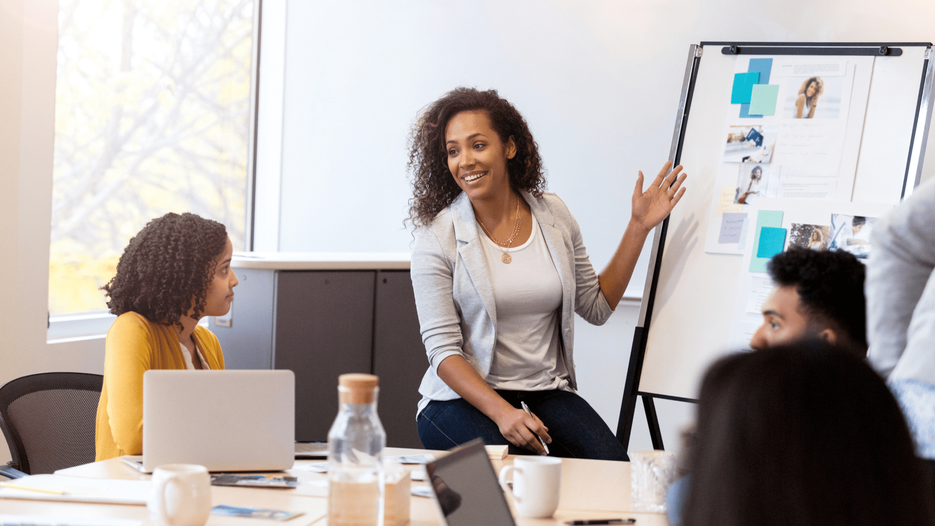 A woman leading a discussion or presentation in a collaborative office setting. She is seated next to a whiteboard covered with notes and images, gesturing while speaking to a small group. Another woman is seated at the table with a laptop, actively listening. The environment suggests an informal team meeting or brainstorming session.