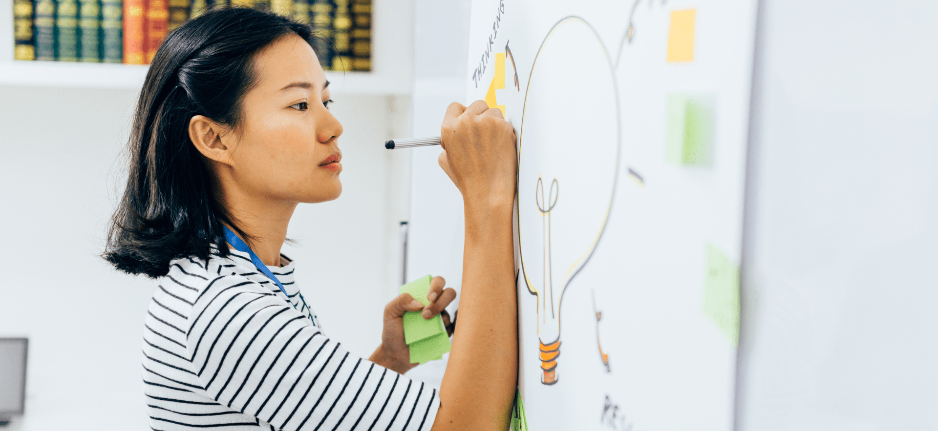 A woman writing on a whiteboard with a diagram of a lightbulb, symbolizing the generation of ideas and creative thinking. The image illustrates the use of visual aids and mnemonic devices in learning and teaching strategies.