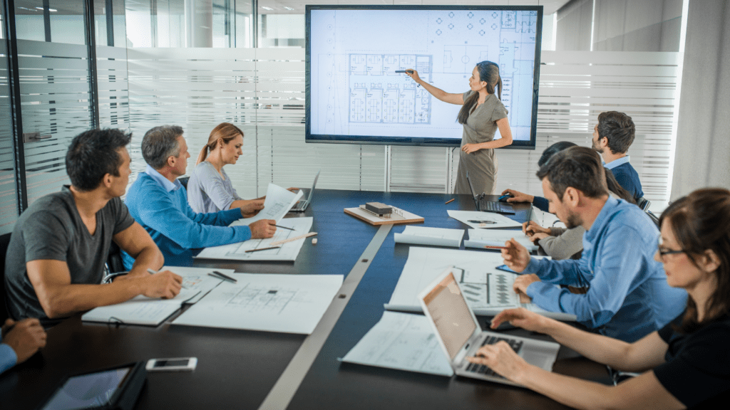 A group of professionals in a conference room attentively listens as a presenter explains architectural plans displayed on a large screen, showcasing a collaborative environment focused on design and strategy.